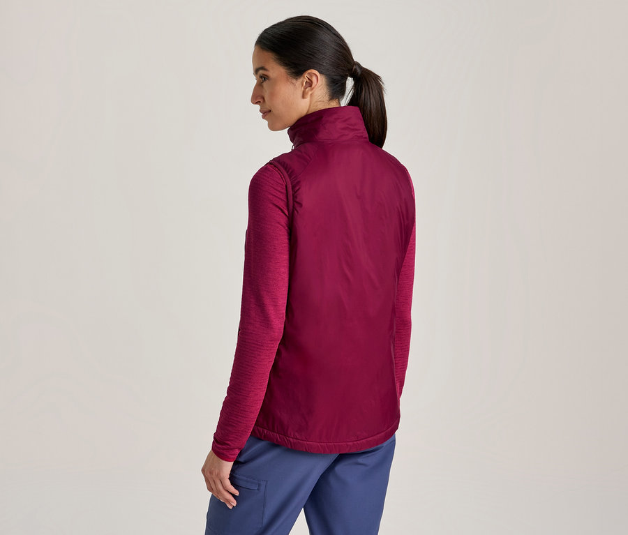 Frau von hinten, leicht gedreht, trägt ein magentafarbenes Langarmshirt, eine dunklere magentafarbene Weste und blaue Hosen. Ihr Haar ist zu einem Pferdeschwanz gebunden.