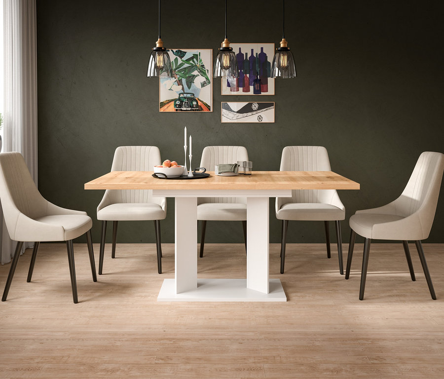 Esszimmer mit hellem Holztisch, weißem Sockel und sechs beigen Stühlen. Auf dem Tisch stehen eine Schale mit Orangen, Kerzen und Gläser. Dunkelgrüne Wand mit drei Bildern und drei Hängelampen. Heller Holzboden.
