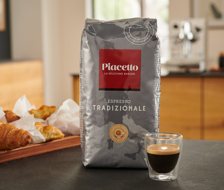 Eine Packung Piacetto Tradizionale Espresso steht auf einem Tisch neben einer Tasse Espresso und einem Tablett mit Croissants.