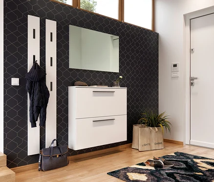 Moderner Flur mit dunkler Tapete und geometrischem Muster. Links ein weißes Garderobenpaneel mit schwarzen Haken, an dem eine dunkle Jacke hängt. Darunter steht eine blaue Tasche. In der Mitte ein weißer schwebender Schuhschrank mit zwei Schubladen und schwarzen Griffen, darüber ein rechteckiger Spiegel. Rechts daneben ein beiger Stoffkorb mit Pflanze und ein Paar Schuhe auf einem gemusterten Teppich vor einer weißen Tür.