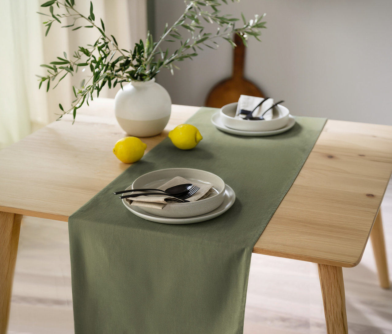 Ein Holztisch ist mit einem khakifarbenen SCHÖNER WOHNEN-Kollektion Tischläufer, zwei Gedecken, zwei Zitronen und einer Vase mit grünen Zweigen gedeckt.