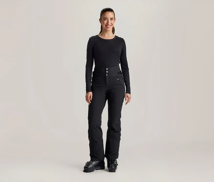 Eine lächelnde Frau steht frontal da und trägt ein schwarzes Langarmshirt, eine schwarze hochgeschnittene Skihose mit Knöpfen und Reißverschlusstaschen sowie schwarze Skischuhe.
