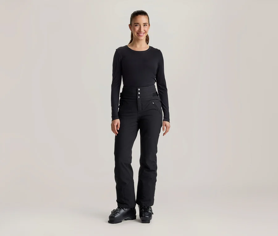 Eine lächelnde Frau steht frontal da und trägt ein schwarzes Langarmshirt, eine schwarze hochgeschnittene Skihose mit Knöpfen und Reißverschlusstaschen sowie schwarze Skischuhe.