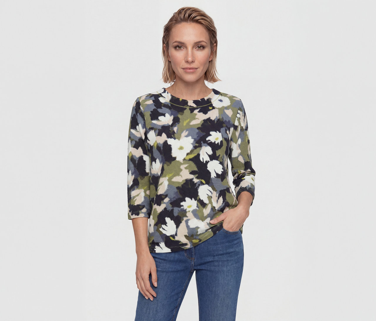 Frau mit kurzen blonden Haaren trägt ein Oberteil mit Blumenmuster in Schwarz, Weiß, Grün, Beige, Grau und blaue Jeans. Eine Hand ist in der Tasche.