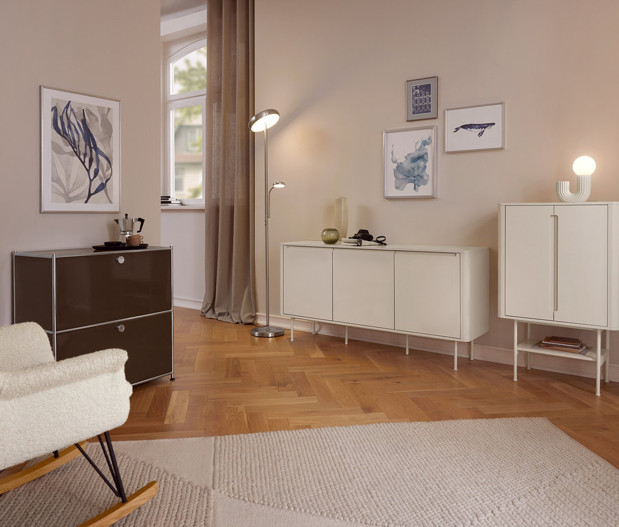 Ein gemütliches Wohnzimmer mit hellen Wänden und Holzboden. Im Vordergrund steht ein cremefarbener Schaukelstuhl neben einem dunkelbraunen Schrank mit einer Mokkakanne. Eine Stehlampe steht zwischen einem langen weißen Sideboard und einem höheren weißen Schrank. Verschiedene gerahmte Kunstwerke schmücken die Wände, und ein heller Teppich bedeckt einen Teil des Bodens.