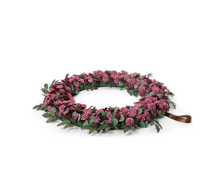Ein runder Blumenkranz mit kleinen rosa und violetten Blüten und grünen Blättern, mit einer braunen Bandschlaufe an einer Seite, vor weißem Hintergrund.