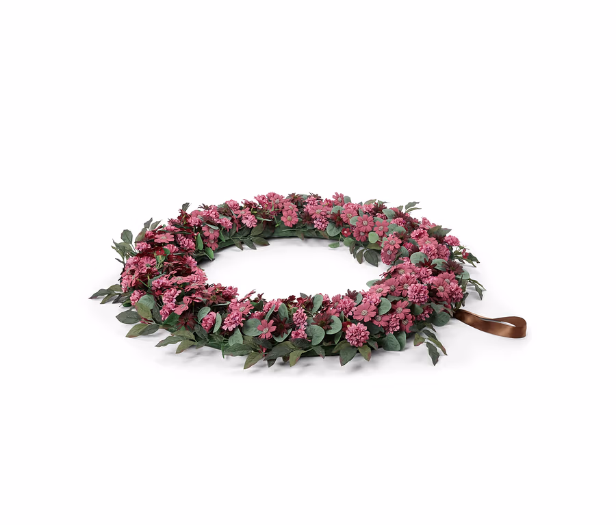 Ein runder Blumenkranz mit kleinen rosa und violetten Blüten und grünen Blättern, mit einer braunen Bandschlaufe an einer Seite, vor weißem Hintergrund.
