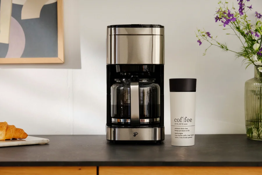 Eine Kaffeemaschine aus Edelstahl und Schwarz mit zwei Glaskannen voller Kaffee steht auf einer dunklen Arbeitsplatte. Daneben befindet sich ein weißer Reisebecher mit schwarzem Deckel, beschriftet mit „cof fee“ und seiner Definition. Links liegt ein Croissant auf einem Marmorbrett, rechts steht eine Vase mit lila Blumen.