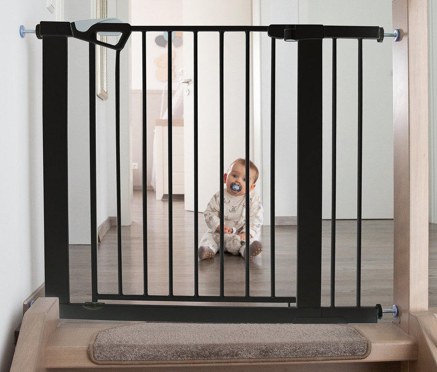 Baby sitzt hinter einem schwarzen roba easySafe+ Tür-und-Treppen-Schutzgitter mit 14-cm-Erweiterung am unteren Ende einer Treppe.