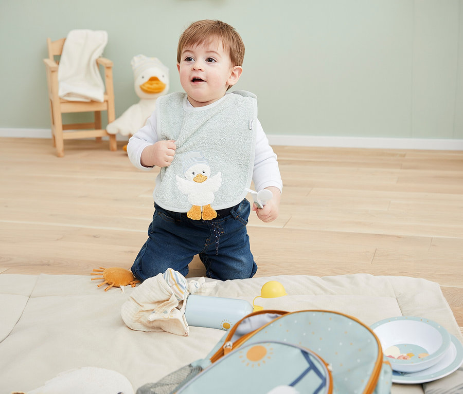 Ein Baby kniet auf einer hellen Matte, trägt ein hellgrünes Lätzchen mit Entenmotiv und hält einen weißen Löffel. Daneben liegen ein hellblauer Rucksack, zwei Teller, eine Flasche und ein kleines orangefarbenes Spielzeug. Im Hintergrund sind ein Holzstuhl und ein großes Enten-Stofftier zu sehen.