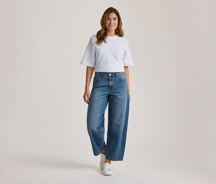 Eine lächelnde Frau steht frontal da und trägt ein weißes T-Shirt mit weiten Ärmeln, blaue weite Jeans und weiße Sneaker.