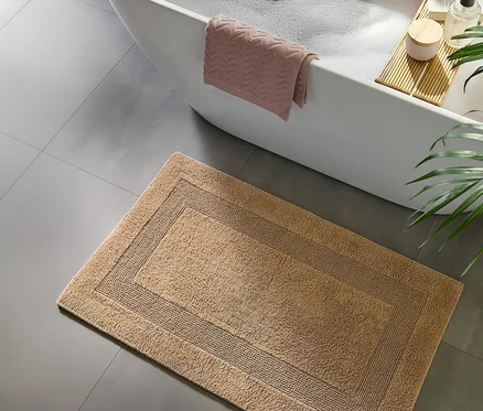 Eine hellbraune Badematte mit dunklerem Rand auf grauem Fliesenboden, vor einer weißen Badewanne mit rosa Handtuch und Holzwannenablage.