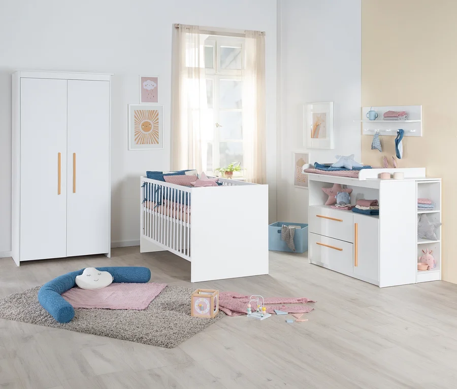 Ein helles Babyzimmer mit weißen Möbeln, darunter ein Kleiderschrank mit Holzgriffen, ein Babybett mit blauer und rosa Bettwäsche und eine Wickelkommode mit Schubladen und Regalen. Auf einem grauen Teppich liegen ein blaues U-förmiges Kissen, eine rosa Decke und Spielzeug.