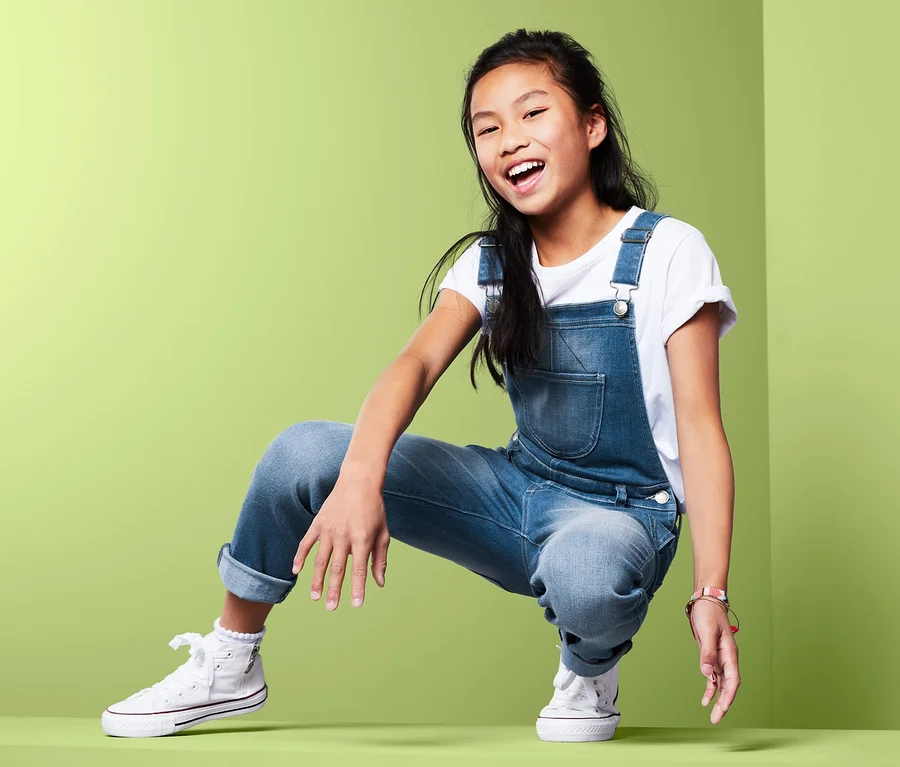 Lächelndes asiatisches Mädchen in Jeans-Latzhose posiert hockend vor grünem Hintergrund.