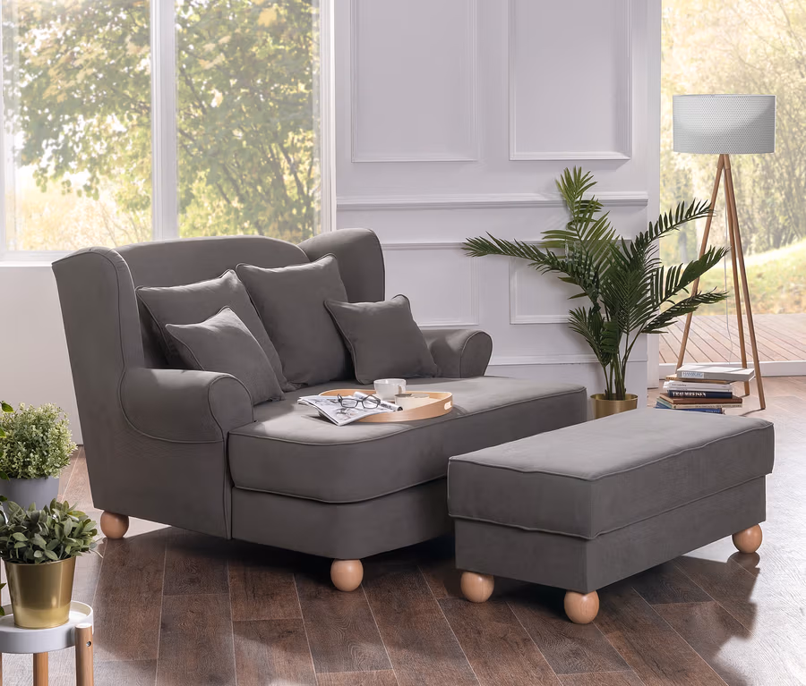Grauer Max Winzer® Polstersessel mit Hockerbank und Kissen in einem Wohnzimmer.