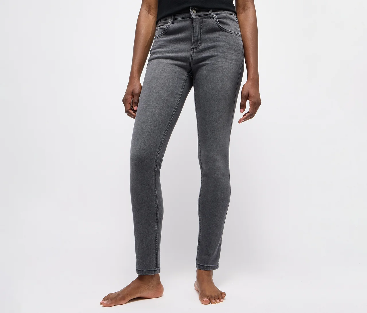 Person trägt ein schwarzes Oberteil und eine dunkelgraue Skinny Jeans, barfuß vor weißem Hintergrund stehend.