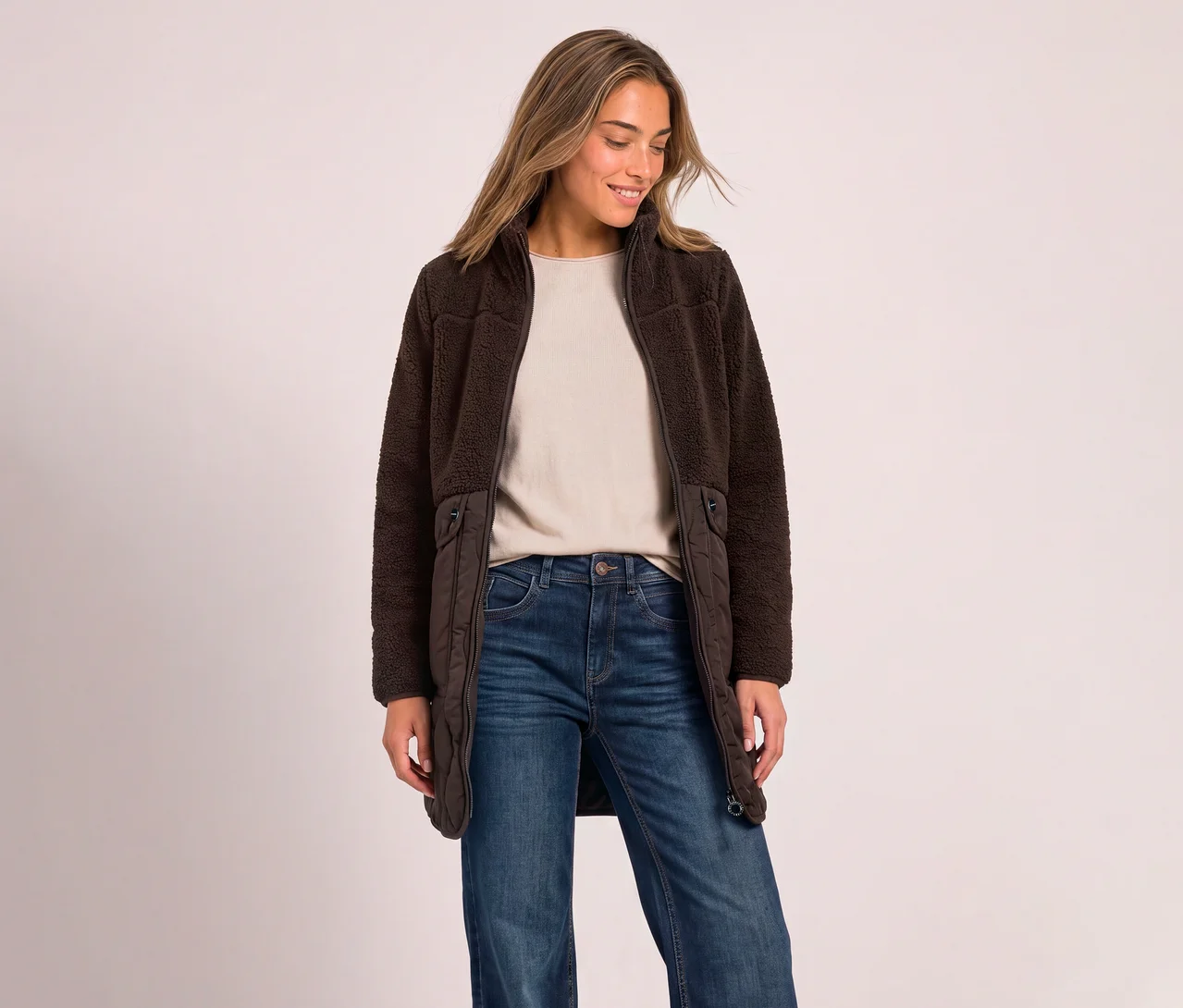 Eine lächelnde Frau mit langen braunen Haaren steht da und blickt nach unten, sie trägt einen braunen Street One Teddymantel über einem hellen Oberteil und blauen Jeans.