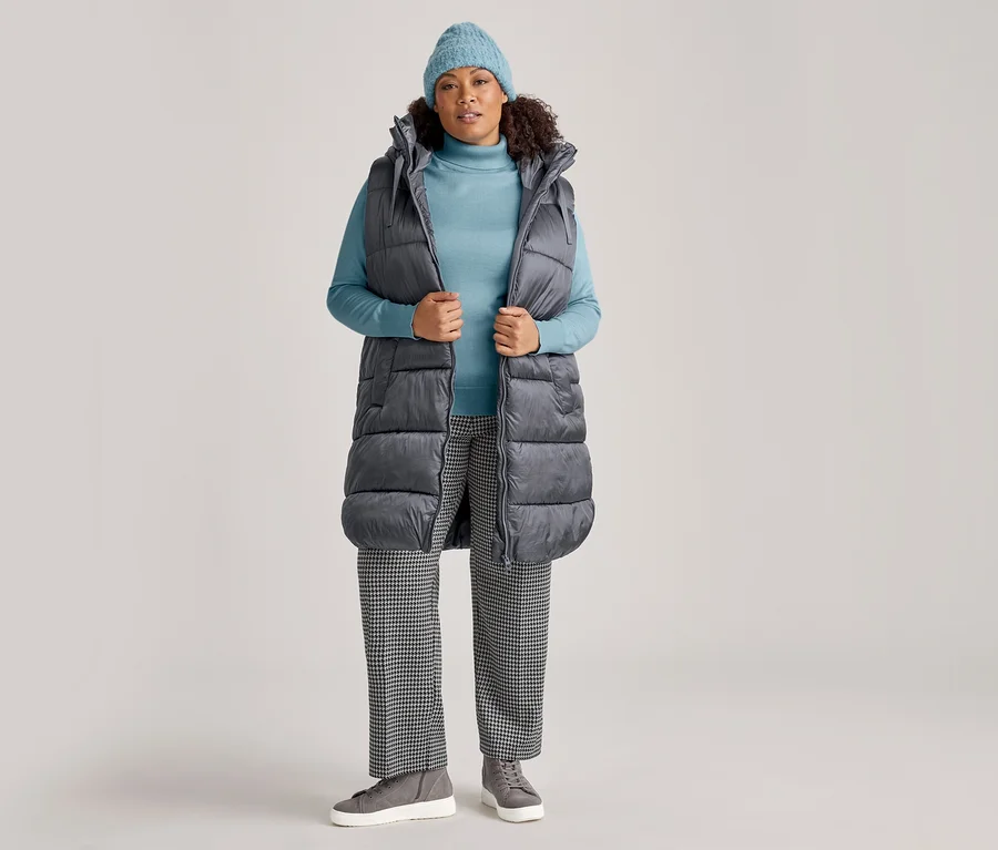 Frau in hellblauer Mütze, hellblauem Rollkragenpullover und langer anthrazitfarbener Steppweste, die sie offen hält. Sie trägt auch schwarz-weiß karierte Hose und graue Sneaker.