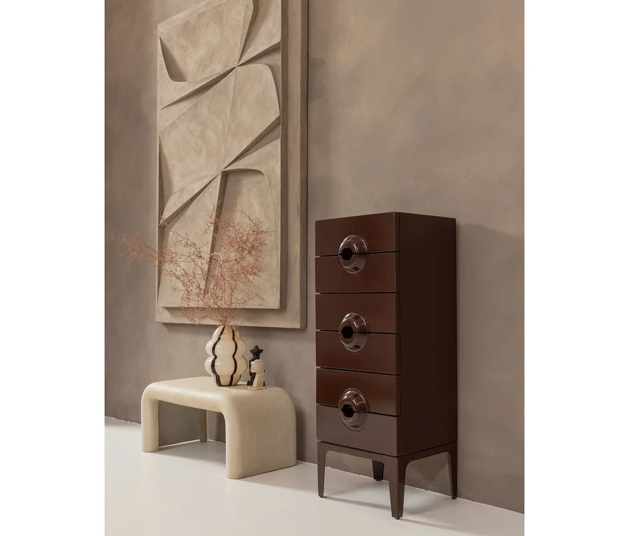 Ein modernes Zimmer mit einer hohen, dunkelbraunen Kommode mit runden Griffen, einer hellen, geschwungenen Bank und einer Vase mit getrockneten Zweigen. Ein großes, strukturiertes abstraktes Kunstwerk hängt an der Wand.