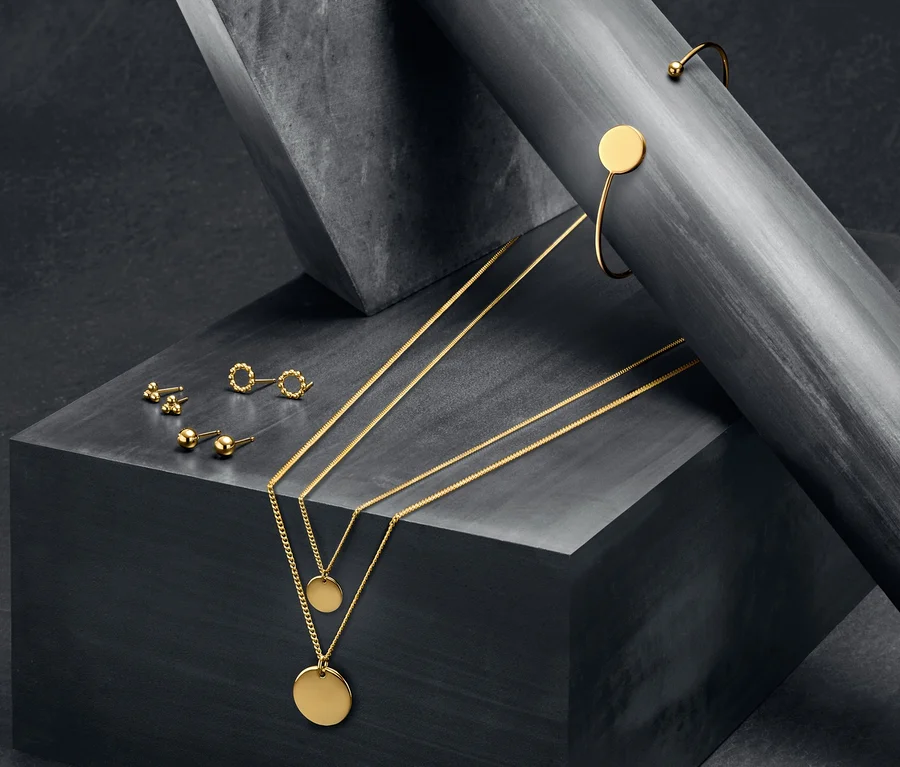 Auf grauem Untergrund liegen Silberschmuckstücke »Pure Collection«: Layer-Silberketten, Silberohrstecker-Sets und ein offener, gelbgoldfarbener Silberarmreif.