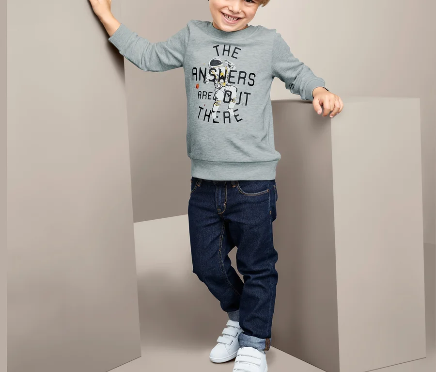 Ein Junge posiert in einem Sweatshirt mit Aufdruck, Jeans und weißen Turnschuhen.