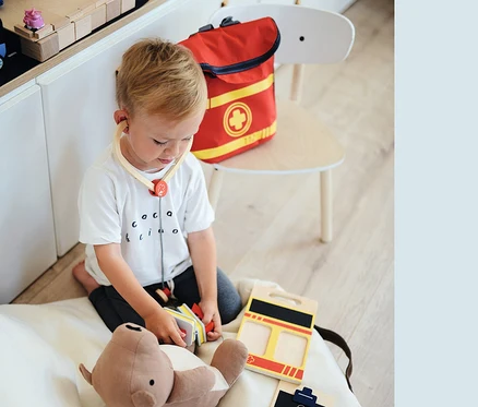 Ein Junge spielt mit einem Teddybär und einem Arztkoffer, trägt ein Stethoskop und daneben liegt ein small foot Notarzt-Rucksack.