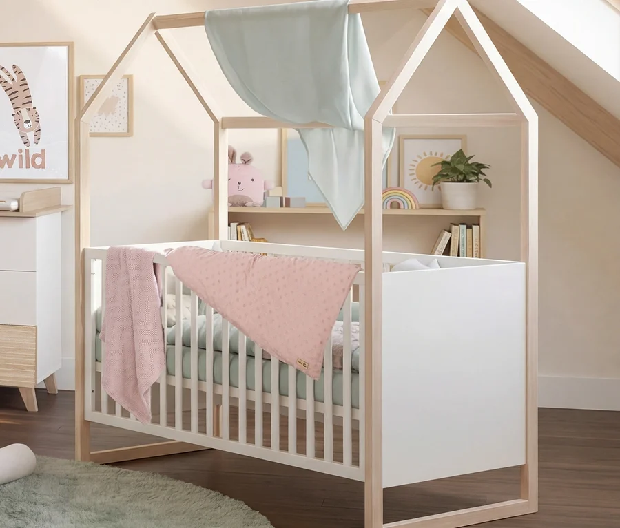 Ein gemütliches Kinderzimmer mit einem hausförmigen Holzbettchen mit weißen Seiten, einer rosa Decke und einem hellgrünen Laken. Ein hellgrüner runder Teppich liegt auf dem dunklen Holzboden. Links steht eine weiße Kommode mit Wickelauflage und einem "wilden" Tigerposter. Rechts befinden sich Regale mit Büchern, einer Pflanze und einer Regenbogen-Dekoration.