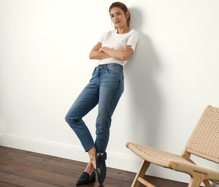 Eine Frau, die sich an eine Wand lehnt, trägt Slimfit-Jeans, ein weißes T-Shirt und schwarze Slipper.