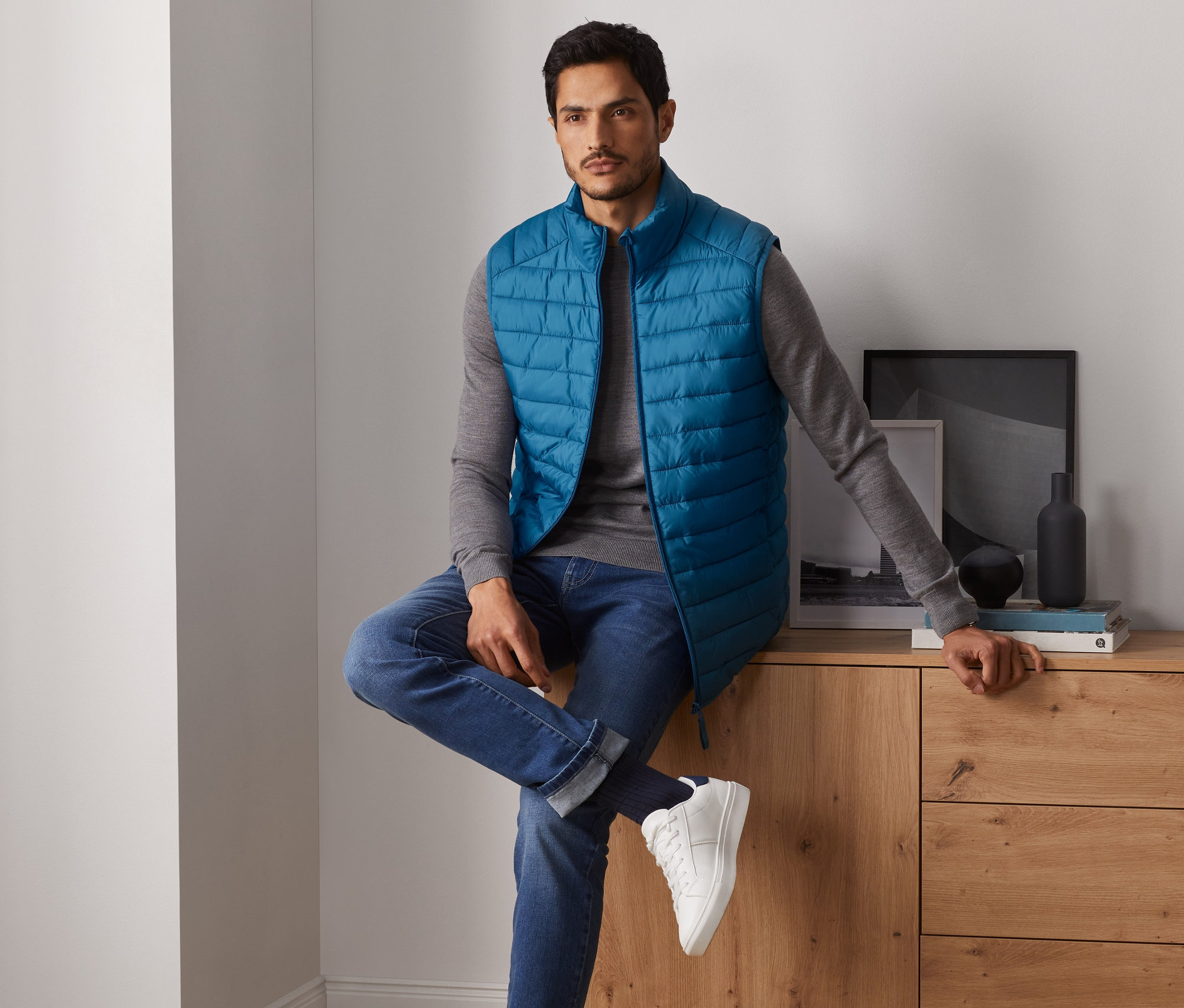 Mann sitzt auf Kommode und trägt Steppweste.