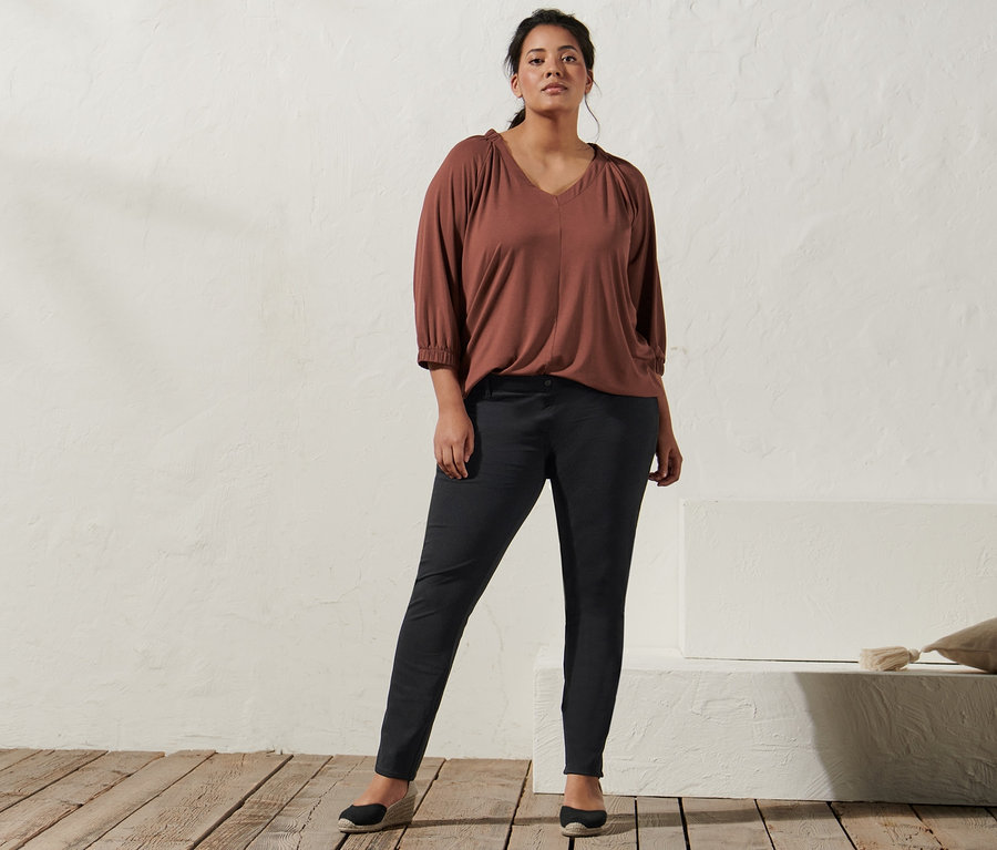 Frau posiert in terrakottafarbenem Shirt mit Raffung, Jeggings und Espadrilles mit Keilabsatz.