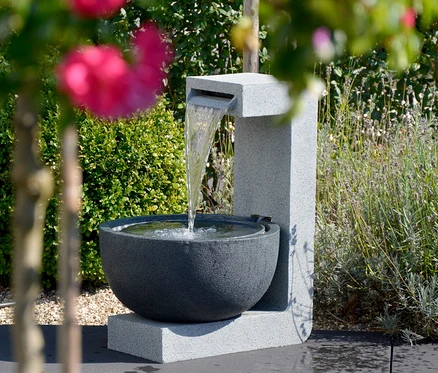 Wasser fließt in die Steinschale des Dobar Gartenbrunnens, im Hintergrund Grün.