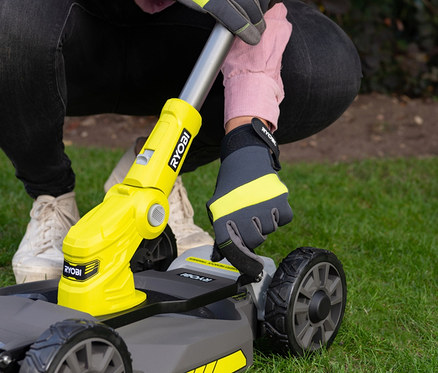Eine Person mit Handschuhen bedient einen RYOBI 18 V One+ Akku-City-Rasenmäher-Set »RY18LMC30A-120« auf Gras.
