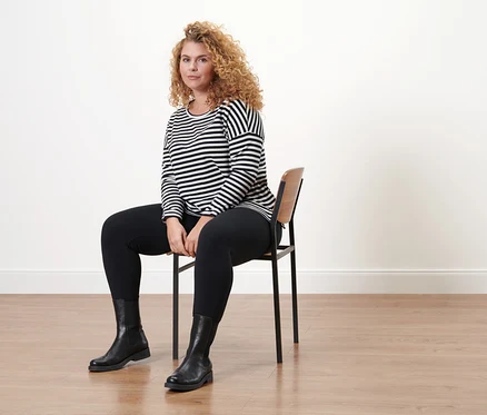 Frau sitzt auf einem Stuhl und trägt Lederboots und ein gestreiftes Langarmshirt.