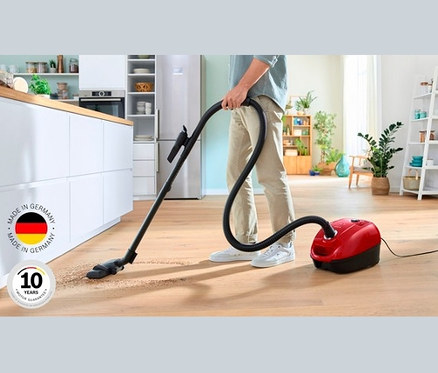 Ein Mann saugt den Boden mit einem roten Staubsauger mit Beutel. Im Hintergrund sind Küchenschränke zu sehen.