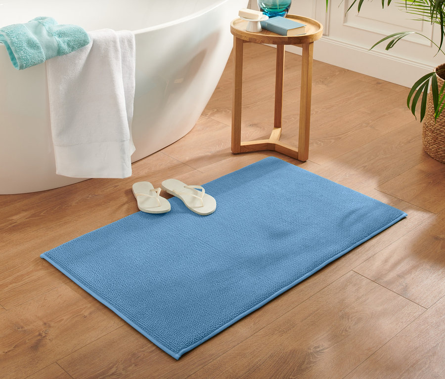 Eine blaue Komfort-Badematte, ca. 60 x 100 cm, liegt auf einem Holzboden neben einer Badewanne. Auf der Matte liegen weiße Flip-Flops.