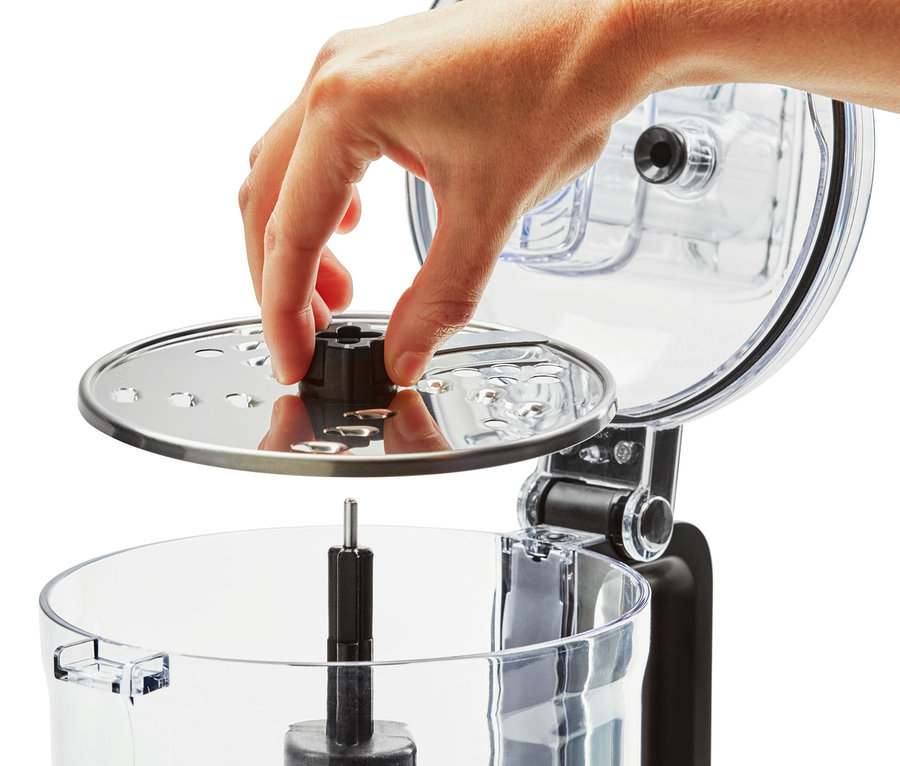 Eine Hand legt eine silberne Scheibe mit Klingen in eine durchsichtige Küchenmaschine des Typs KitchenAid Zerkleinerer »5KFP0719«.