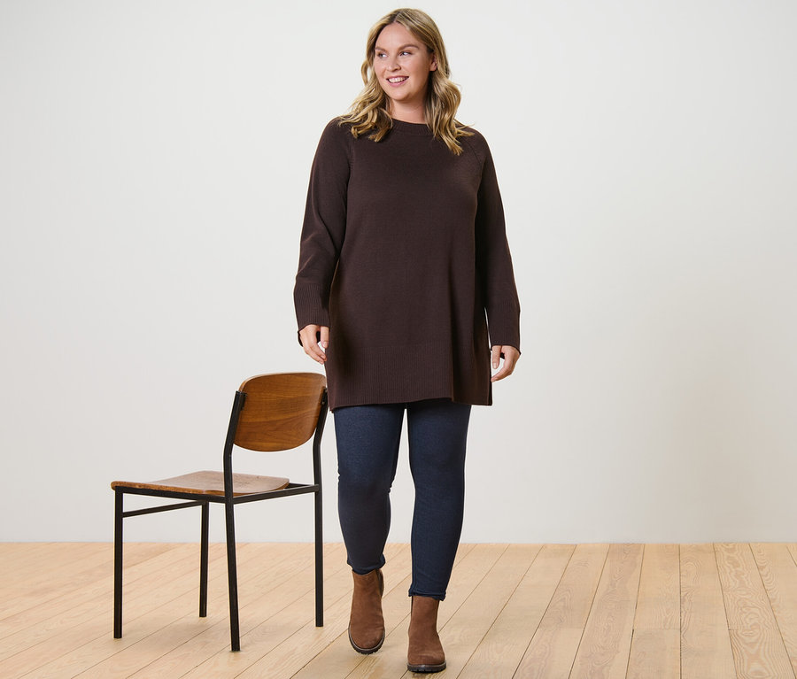 Eine Frau trägt einen langen, dunkelbraunen Strickpullover und gefütterte Leggings in Jeansoptik und geht.