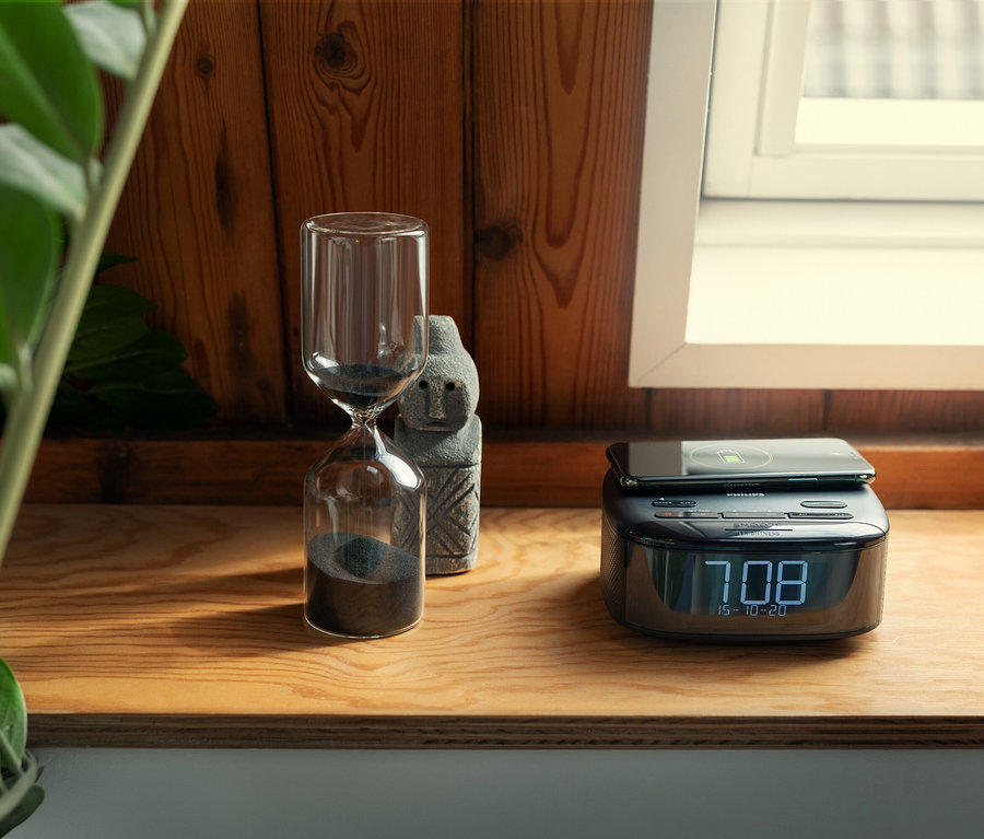 Tischplatte mit Sanduhr, Steinstatue und schwarzem Philips DAB-Radiowecker TAR7705 mit Handyladefunktion.