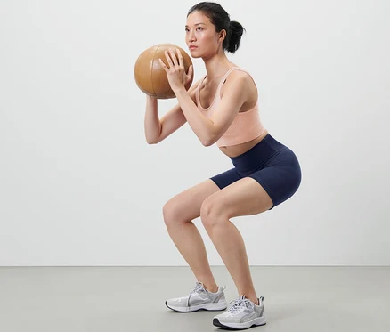 Frau macht Kniebeugen mit Medizinball und trägt kurze Sporttight.