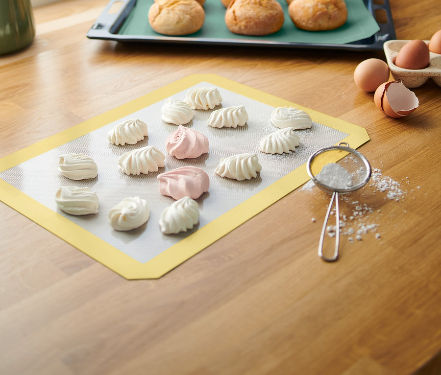 Auf einer Holzfläche liegen ein gelbe Backmatte mit weißen und rosa Baiser. Im Hintergrund befindet sich ein Backblech mit gebackenem Brot.