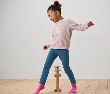 Ein Mädchen in einem rosa Einhorn-Sweatshirt und blauen »Mia« Kinder-Jeggings spielt mit Holzbausteinen.