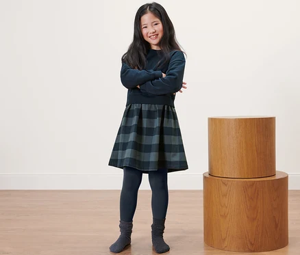 Ein lächelndes Mädchen steht mit verschränkten Armen da und trägt Kinder-Thermoleggings, ein Kleid und einen Pullover.