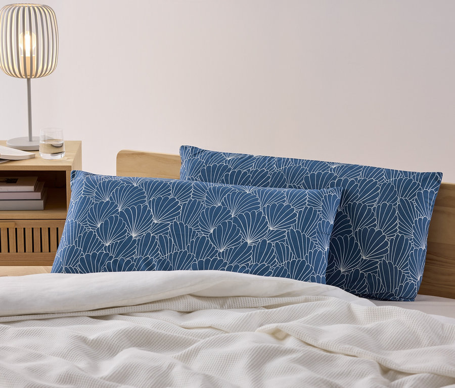 Zwei blaue Microfaser-Kopfkissenbezüge mit weißem Muschelmuster liegen auf einem Bett neben einem Nachttisch mit Lampe.