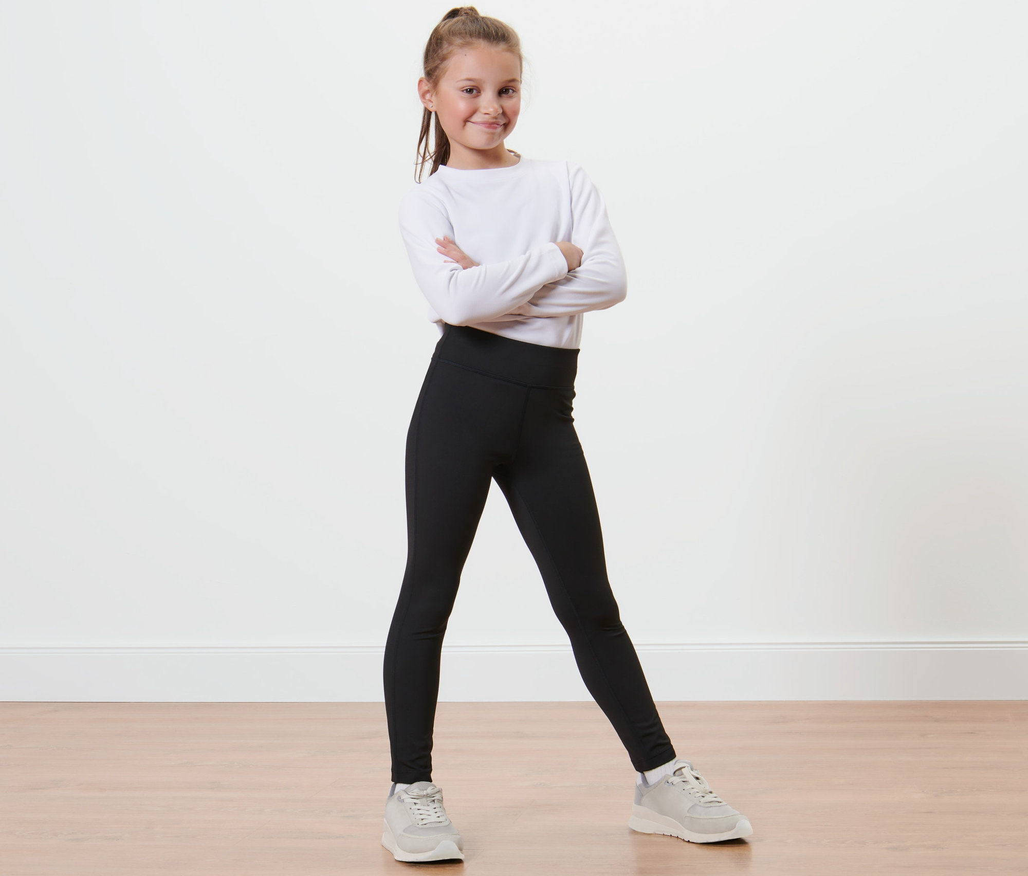 Lächelndes Mädchen posiert mit verschränkten Armen in Kinder-Sporttight, langärmligem, weißen Shirt und Turnschuhen.