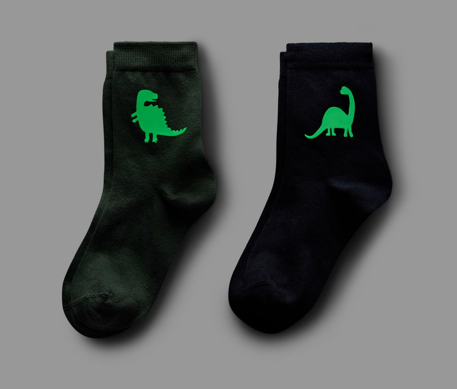 Zwei Paar Socken, eines dunkelgrün mit Dinosaurier-Motiv, eines schwarz mit Dinosaurier-Motiv, beide Paare 'Glow in the Dark'.