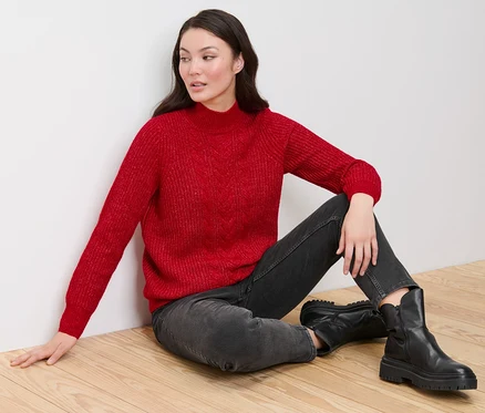 Eine Frau sitzt auf einem Holzboden und trägt einen roten Grobstrickpullover mit Zopfmuster, schwarze Jeans und schwarze Stiefel.