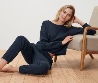 Blonde Frau sitzt auf dem Boden neben einem Sessel und trägt einen dunkelblauen Hausanzug.
