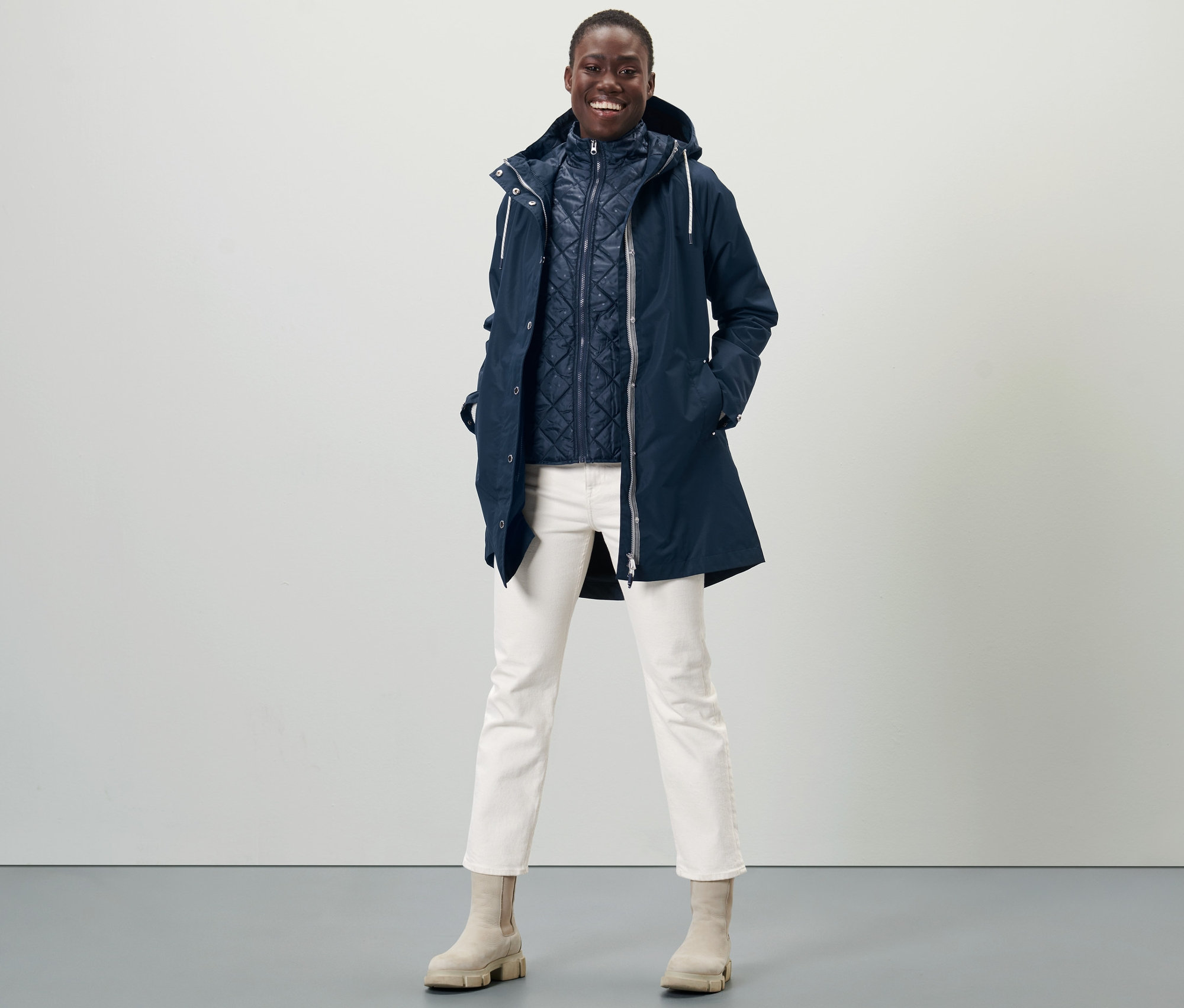Frau posiert in 3-in-1-Regenjacke, weißen Hosen und beigen Stiefeln.