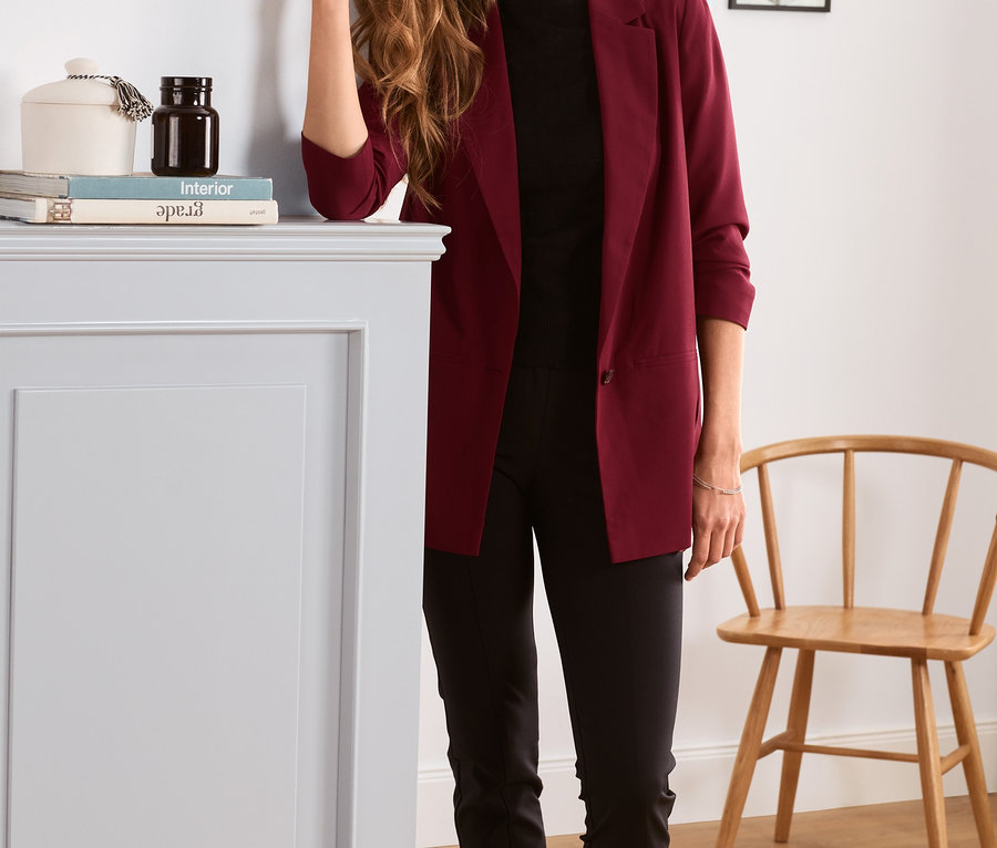 Frau posiert in einem bordeauxroten Blazer mit gerafften Ärmeln.