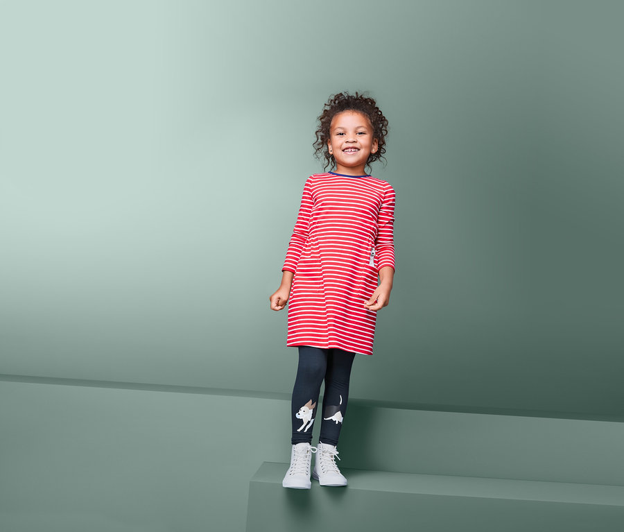 Lächelndes Mädchen in rot-weiß gestreiftem Langarmkleid und dunkelblauen Leggings mit Hundemotiv steht vor grünem Hintergrund. 2 Leggings.